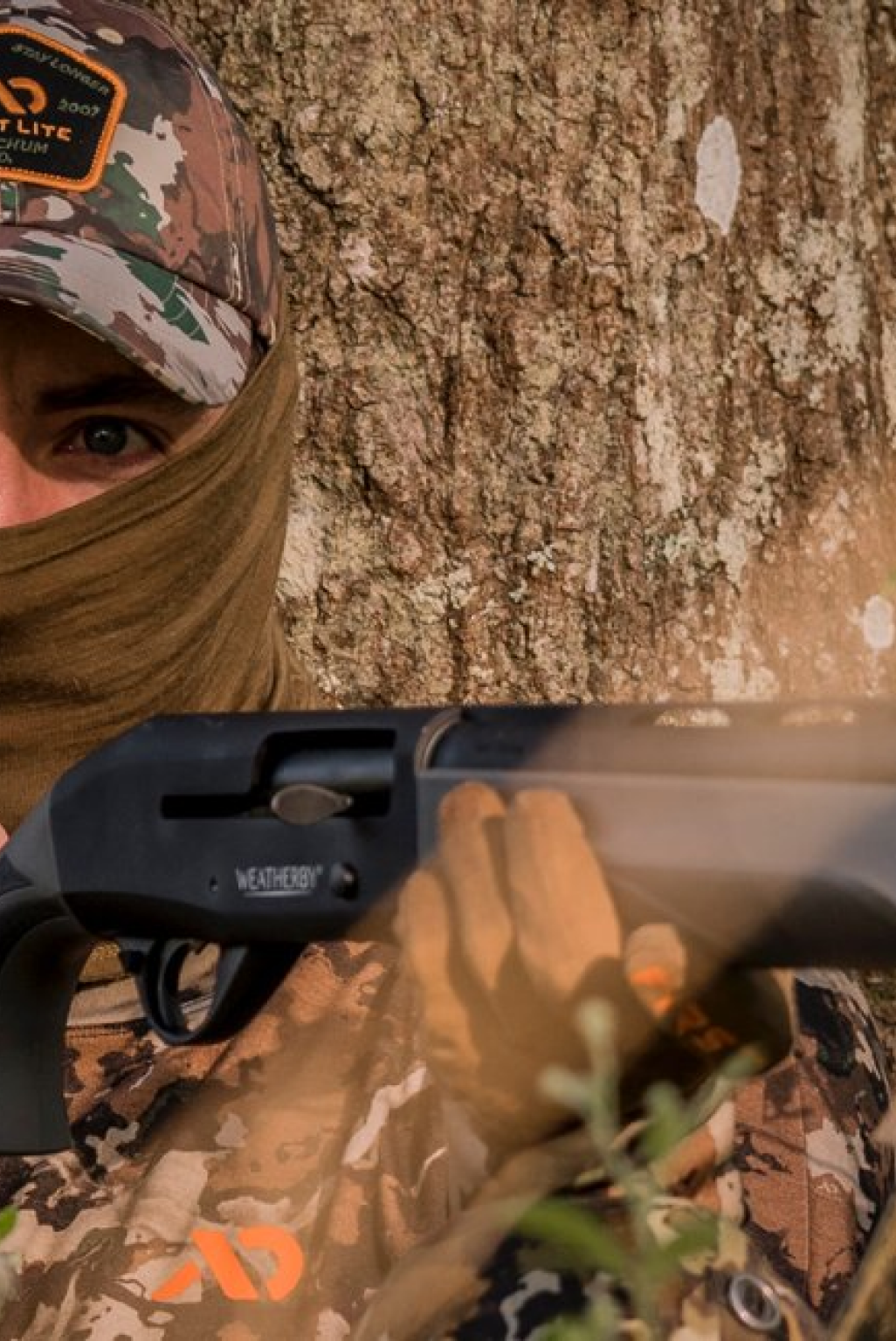 Person in camo holds a black Weatherby shotgun, eyes visible, against tree bark.