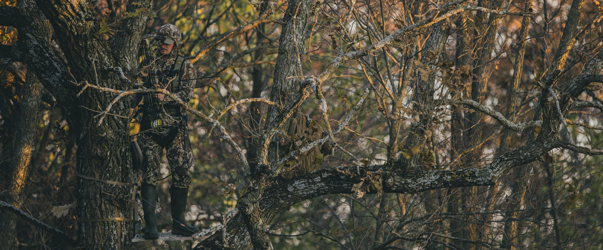 Green camo hunter holds bow on tree stand in a forest.