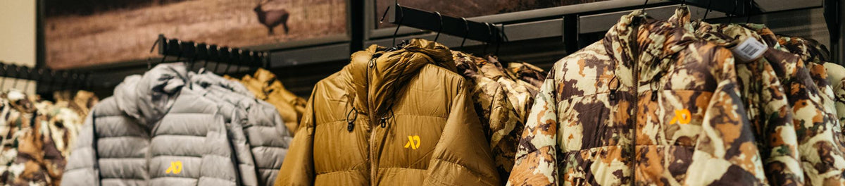 Row of grey, brown, and camouflage puffer jackets with a yellow logo on a rack.