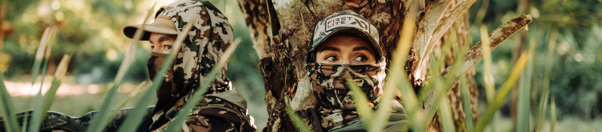 Two people in camo gear, wearing 'First Lite' cap and face masks, blending in foliage.