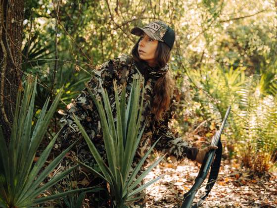 Hunter in green/brown camo gear, cap, gloves, holding a rifle, looking left in dense woods.