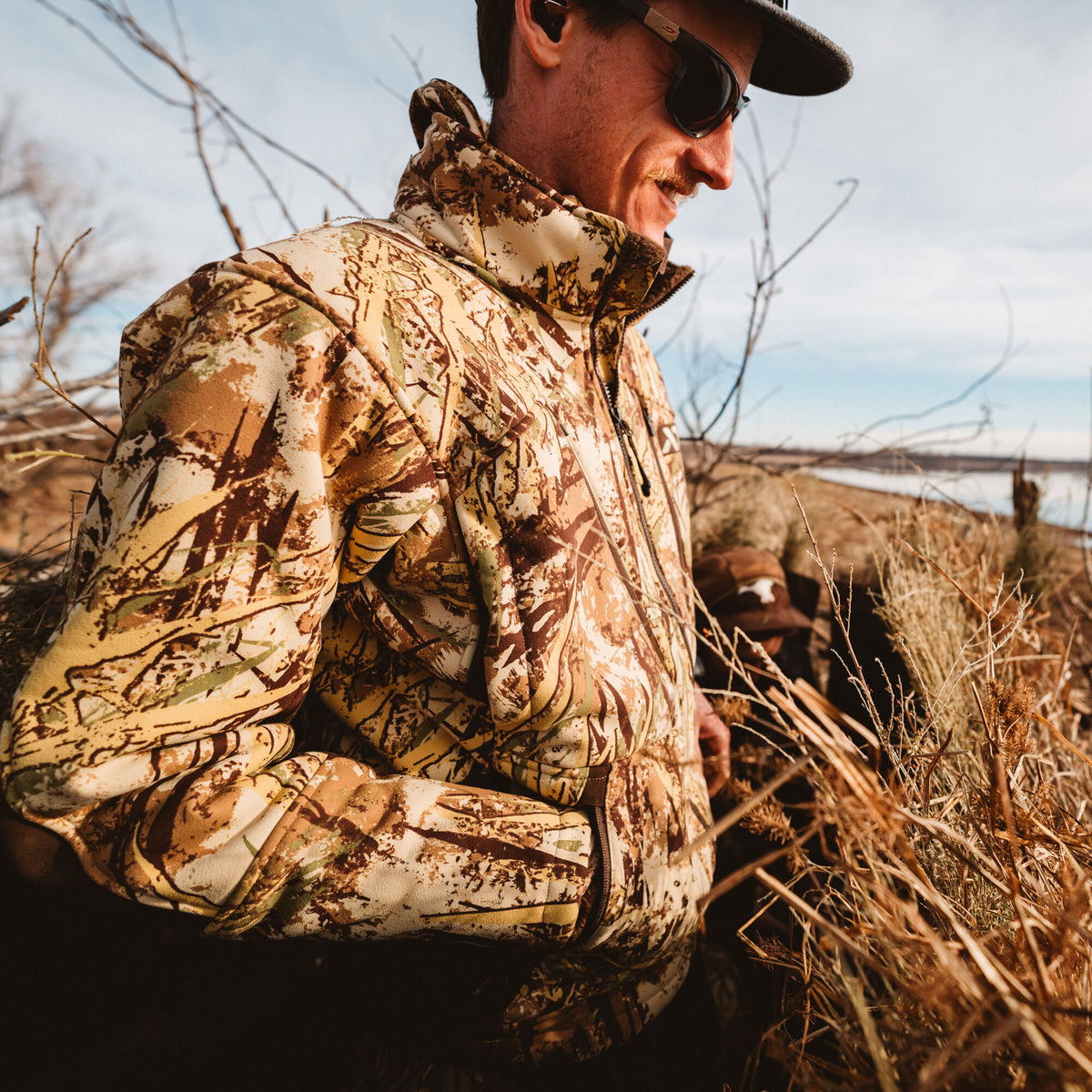 A man wears a multi-toned brown, tan, and green camouflage insulated jacket with a full zipper outdoors.