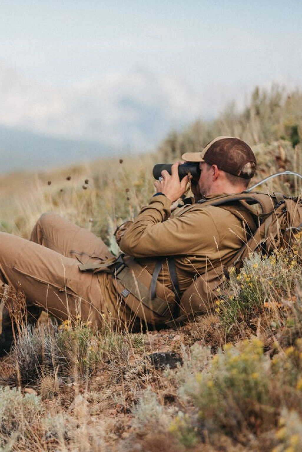 Coyote brown long-sleeve shirt & pants on man using binoculars in tall grass.