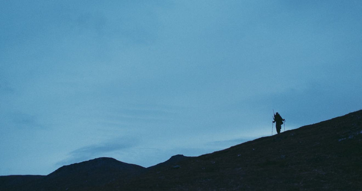 Dark silhouette of a hiker with trekking poles climbing a steep hill under a blue sky.