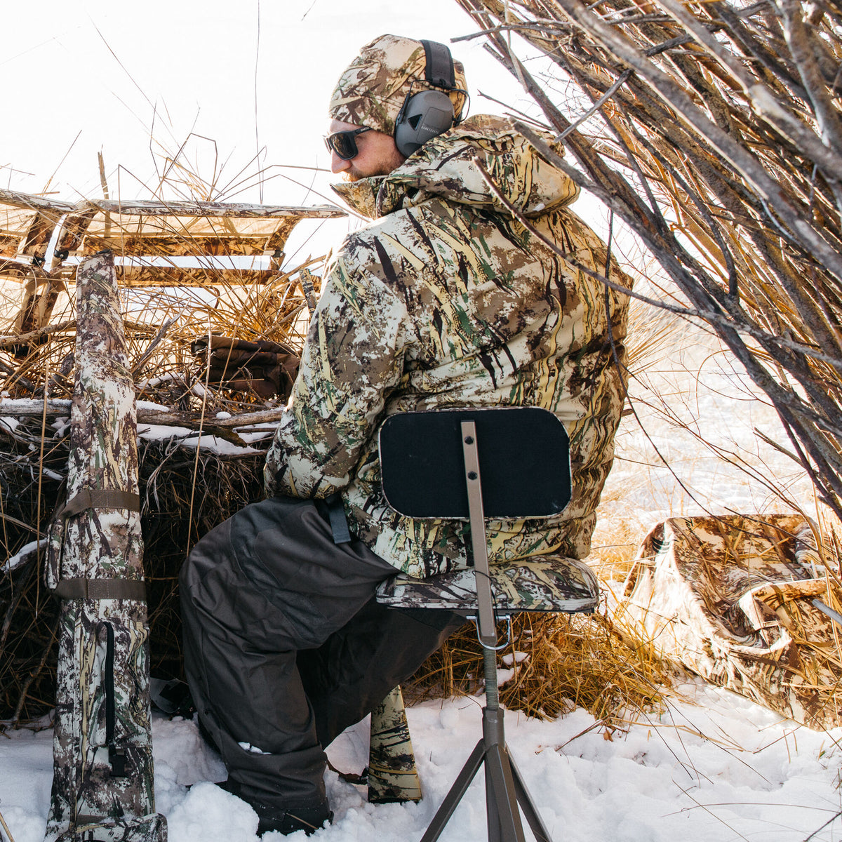 A hunter wearing a multi-pattern camouflage jacket, beanie, and ear protection sits on a dark adjustable hunting stool beside a camouflaged rifle case in a snowy blind.