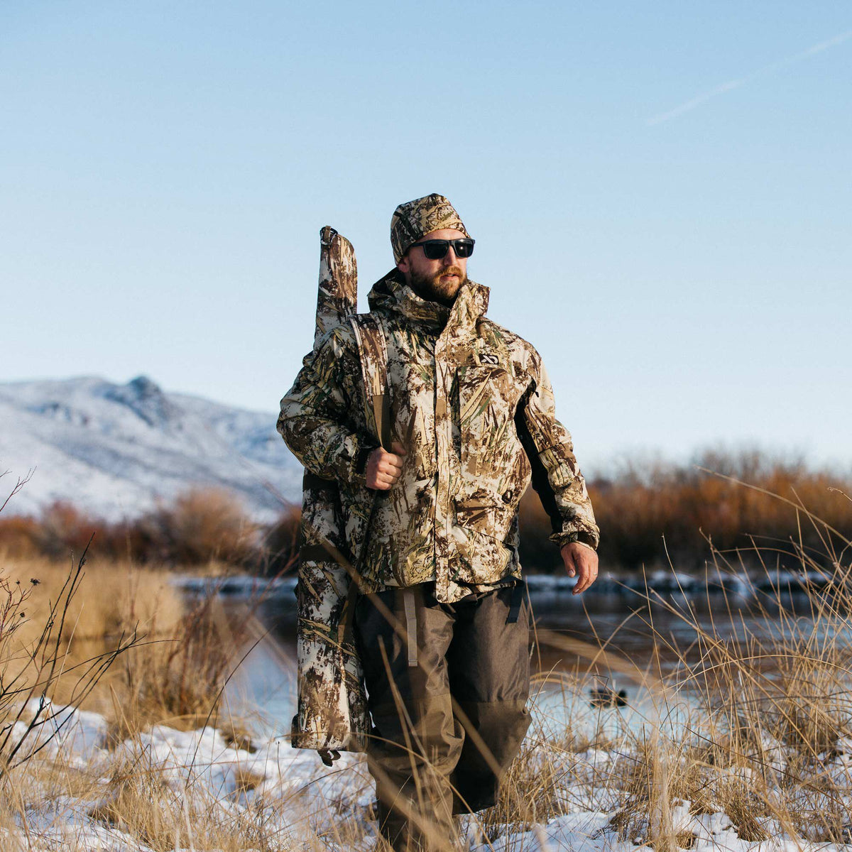 A man wearing a wetlands camouflage hunting jacket, matching hat, and dark waders carries a matching gun case through a snowy marsh.