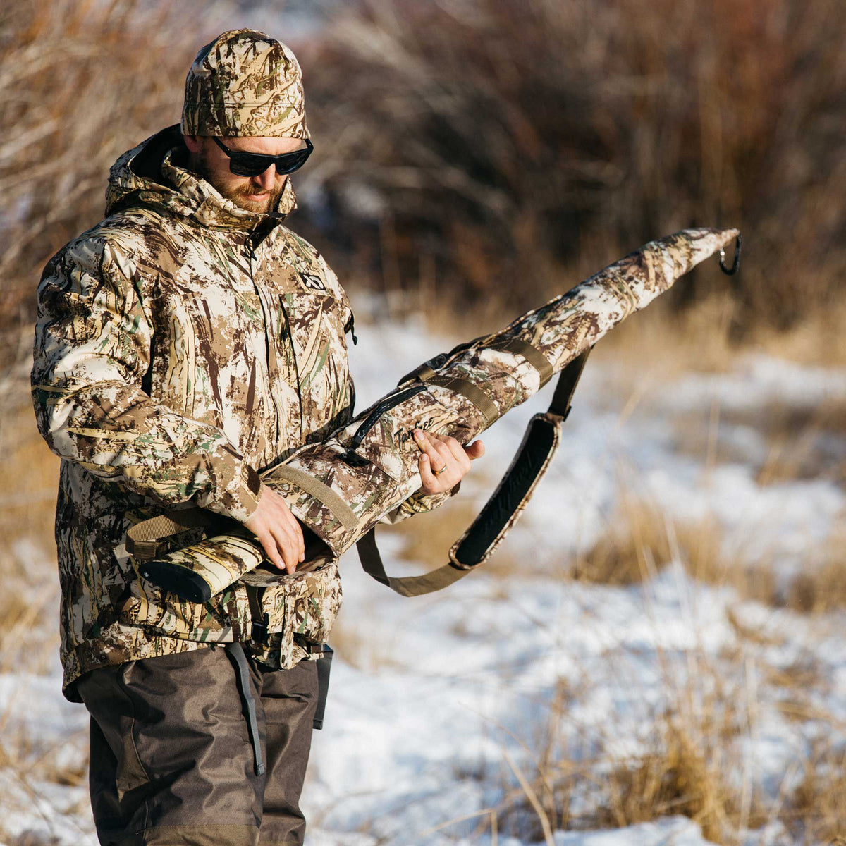 A man in multi-color camouflage hunting jacket, beanie, and pants holds a matching padded rifle case with a shoulder strap and Tanglefree logo.