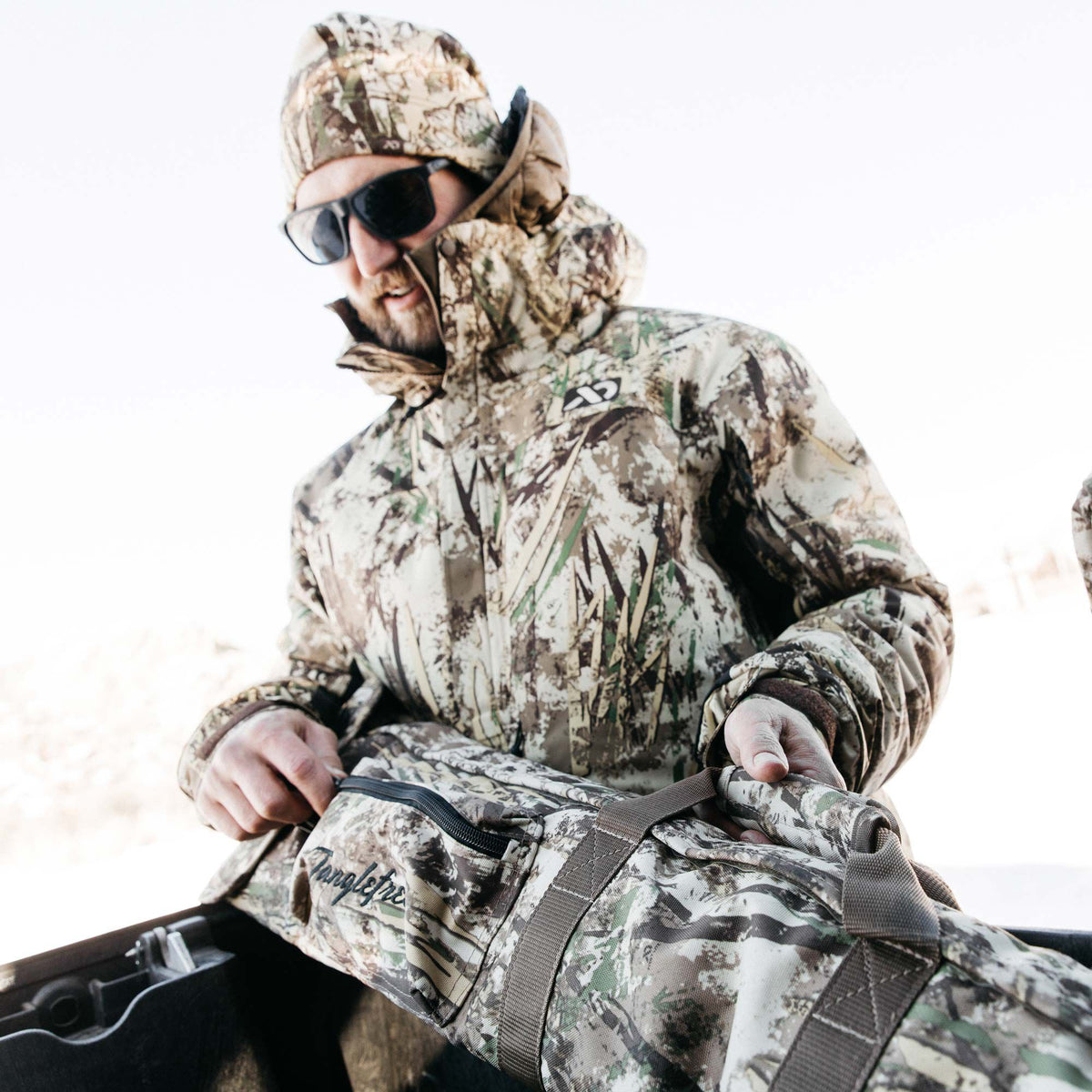 Camouflage duffel bag with a visible zipper and brown carry straps, featuring the "Tanglefree" logo, held by a person wearing matching camo.