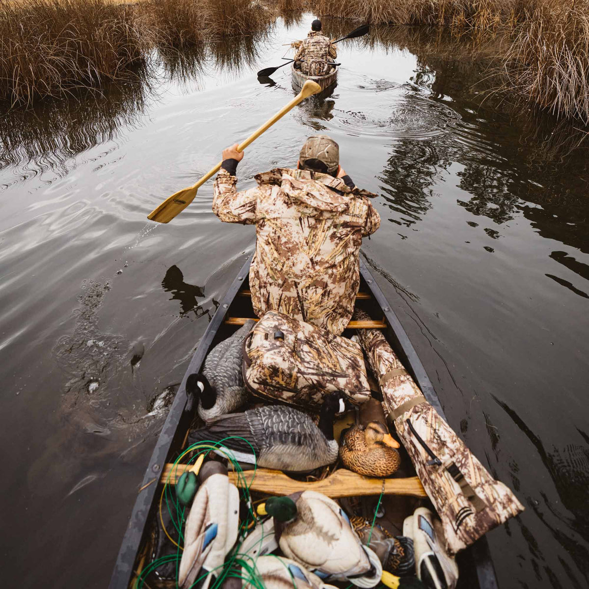 Two dark canoes, one laden with various colored duck and goose decoys and camouflage gear, paddle through a marsh.