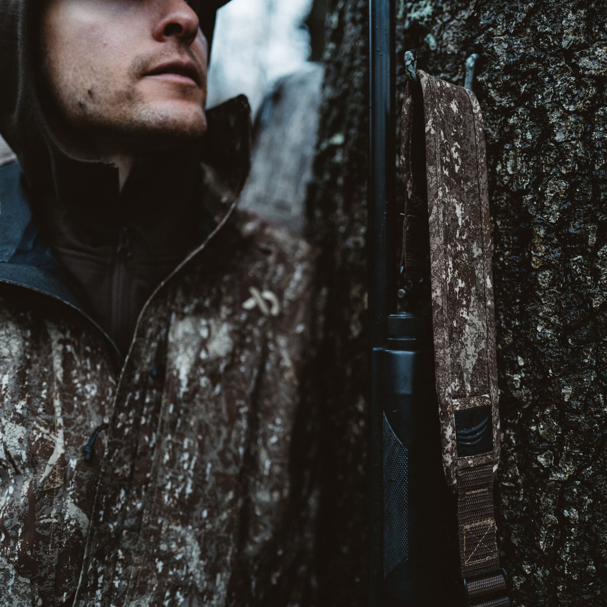 Close-up of a person in a brown camouflage jacket beside a black rifle with a patterned brown camouflage sling hanging vertically on tree bark.