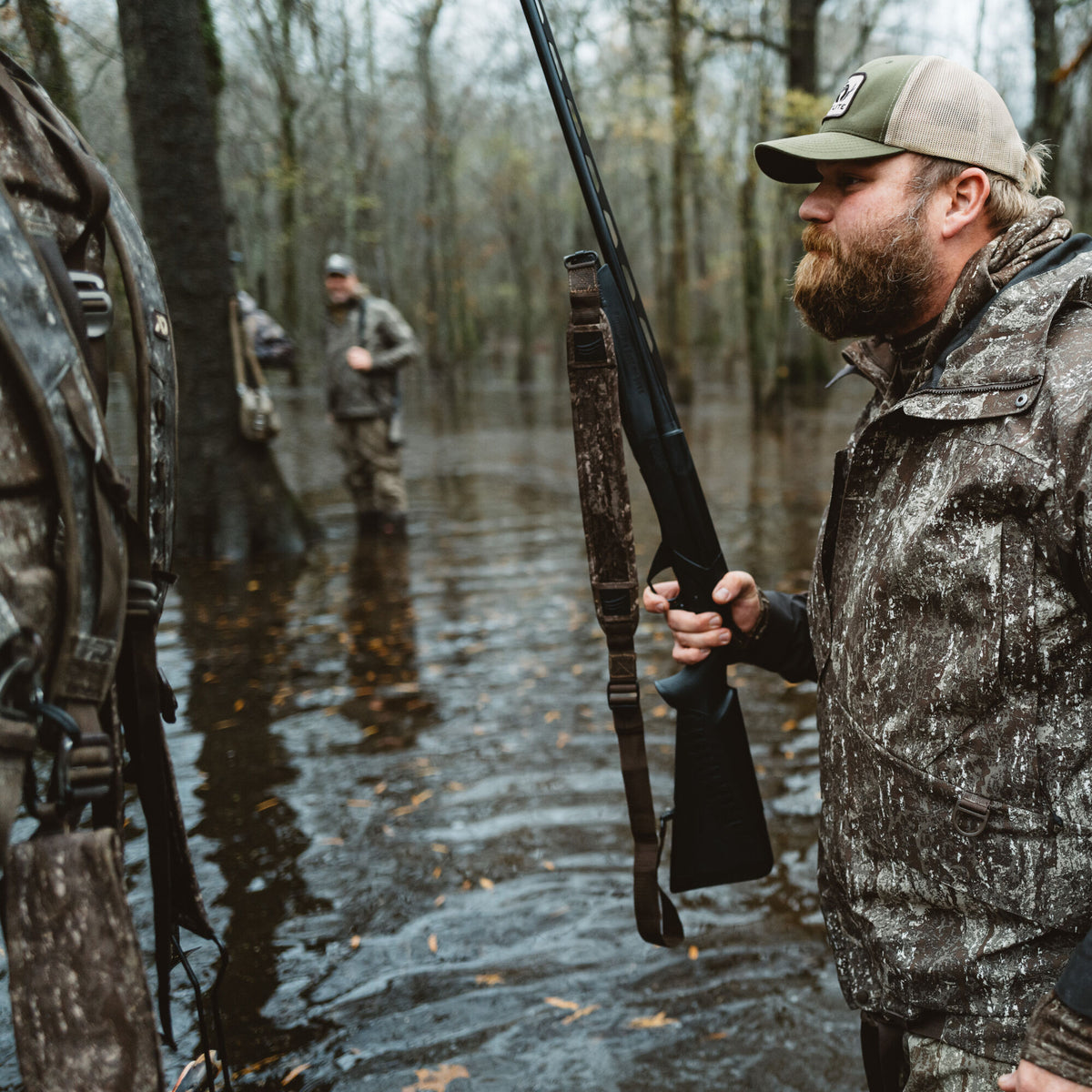 A man in a green and tan trucker hat and brown/white speckled camouflage hunting jacket holds a black shotgun in a flooded forest.