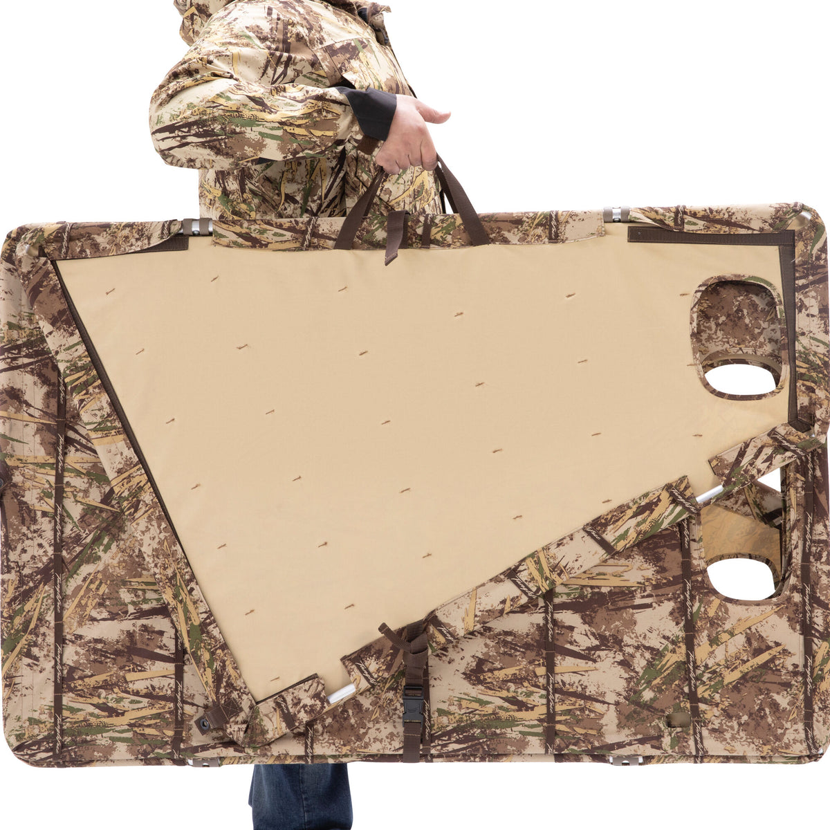 A partially unfolded camouflage hunting blind with a tan interior, two viewing ports, and brown carry straps, is held by a person in matching camouflage.