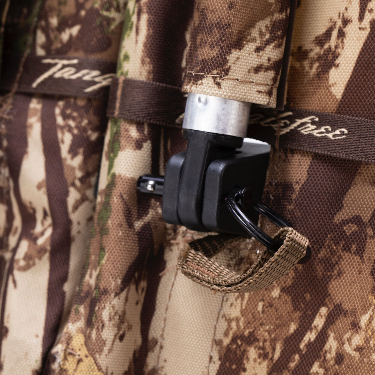 Close-up of a brown and tan camouflage fabric item with a silver and black quick-release attachment, a tan woven loop, and a 'Tanglefree' label.