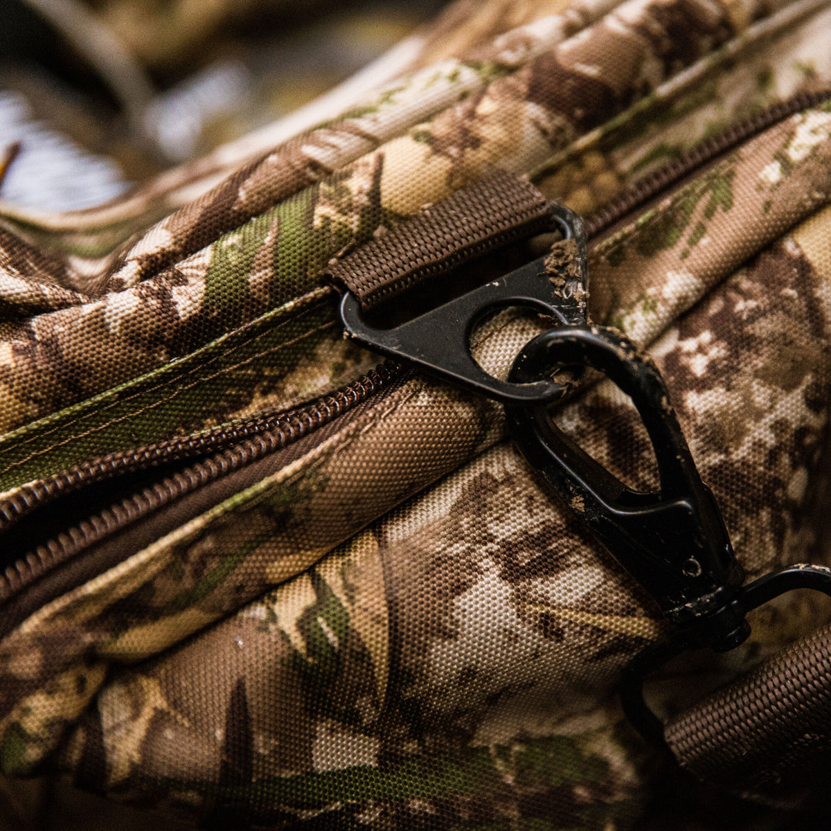 A close-up of a camouflage tactical bag with a brown zipper and a black metal swivel clip on its strap.