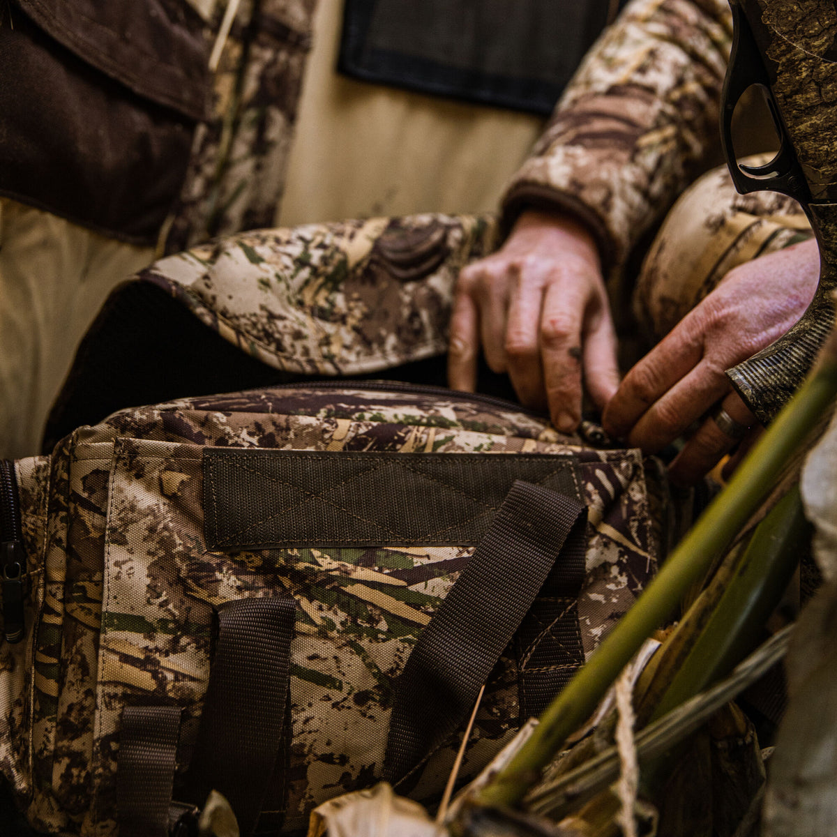 A person's hands unzip a camouflage hunting pack featuring dark brown webbing straps, with a camouflaged shotgun stock visible on the right.