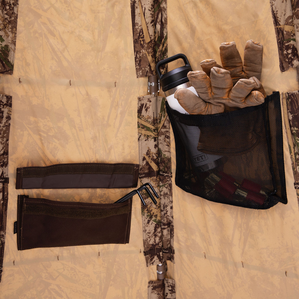 Inside a tan camouflage hunting blind, showing brown utility pockets holding stakes and a mesh pocket with a white bottle, gloves, and shotgun shells.