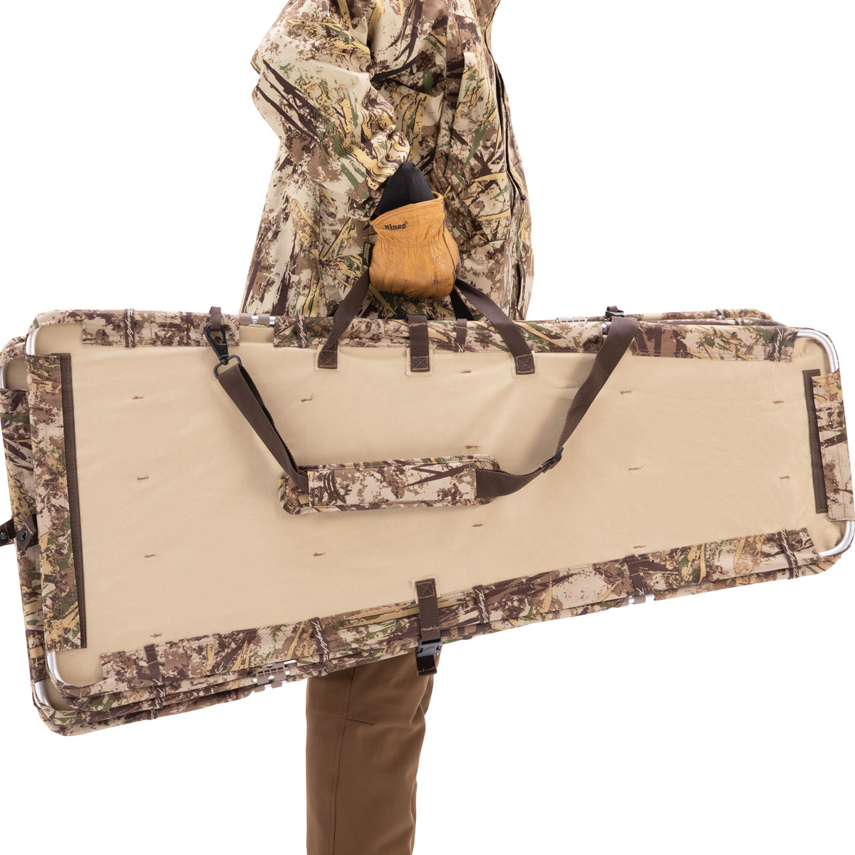 A person in camouflage carries a folded hunting blind with a camouflage exterior, beige interior, metal frame, and carrying straps.