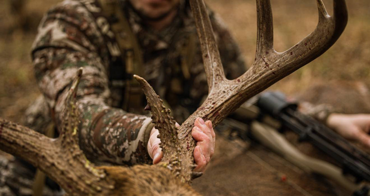 Camo-clad hunter holds brown, multi-tined deer antlers with blood spots; bow nearby.
