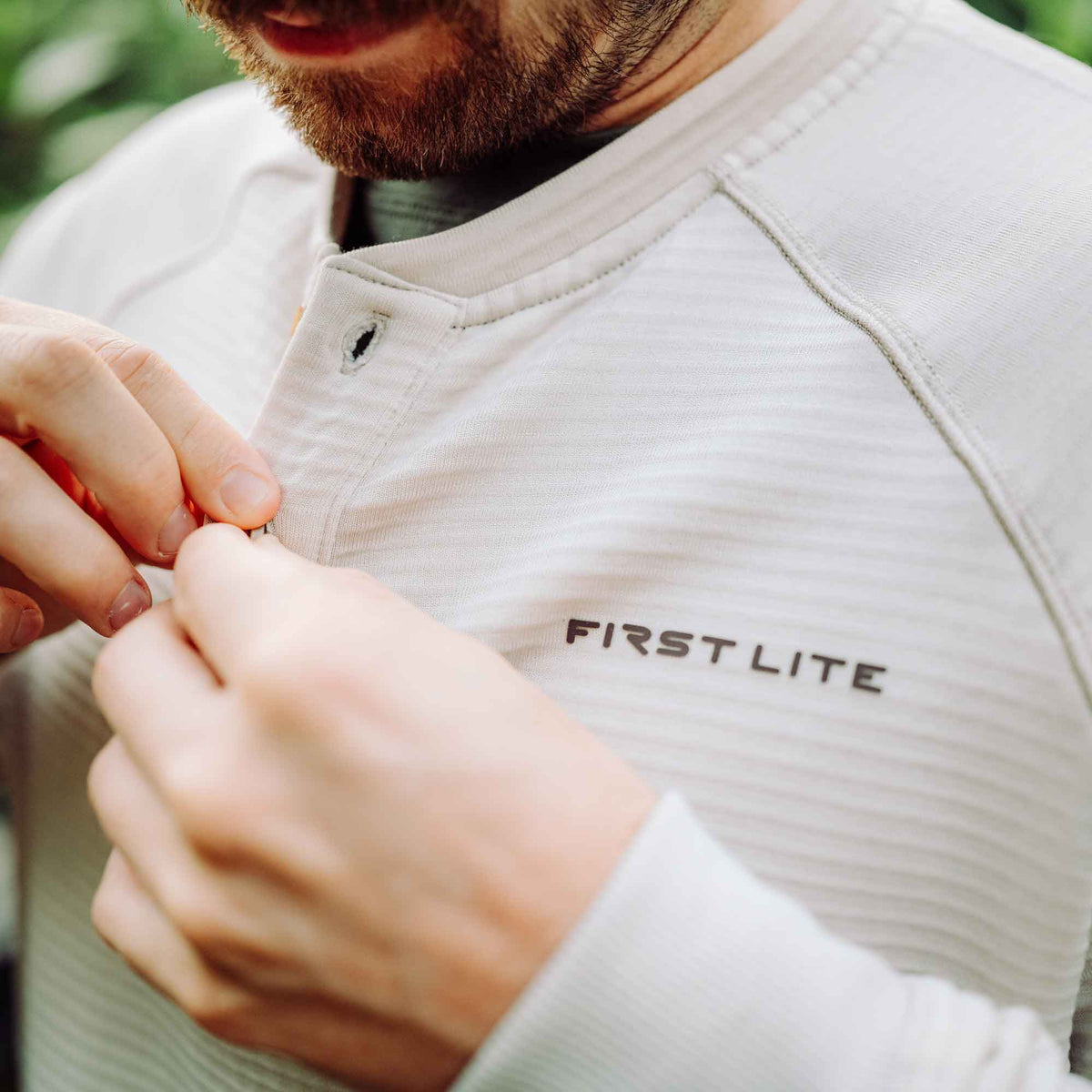 A man buttons a light tan ribbed long-sleeve henley shirt with a "FIRST LITE" logo on the chest.