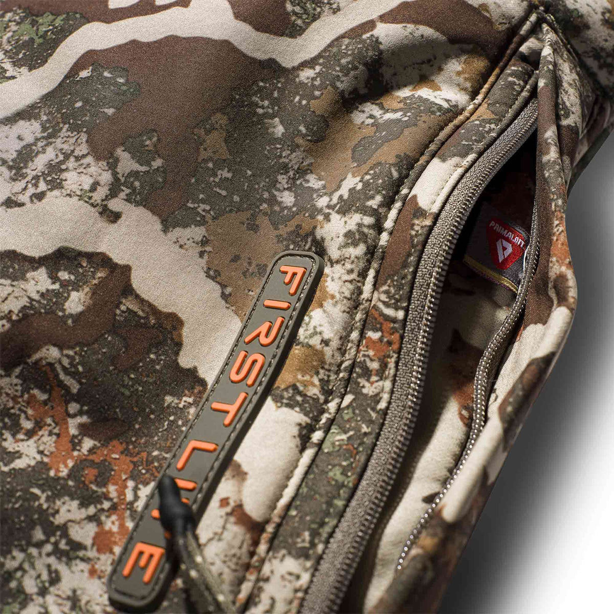 Close-up of a camouflage jacket with an open zippered pocket showing a Primaloft tag and an orange First Lite zipper pull.