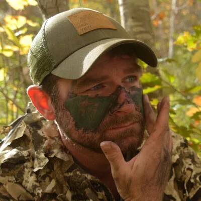 Olive green trucker cap with a tan leather patch, worn by a man with camo paint.