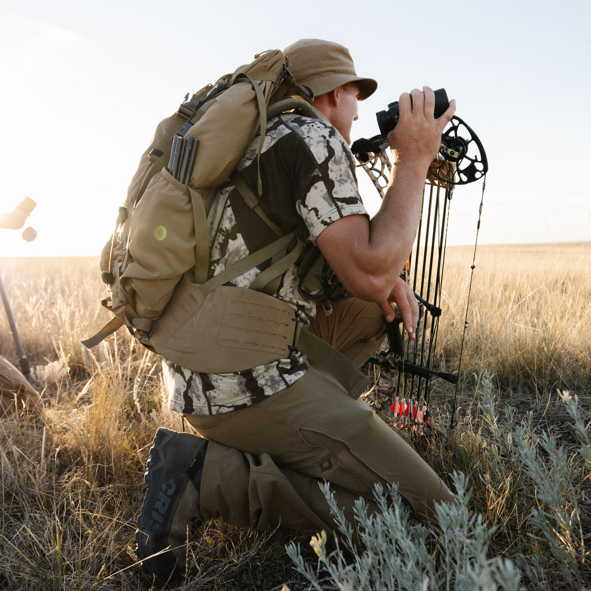 A man wearing a tan tactical backpack with a MOLLE hip belt, tan bucket hat, camo shirt, and tan pants kneels in grass, looking through black binoculars while holding a compound bow with red-fletched arrows.