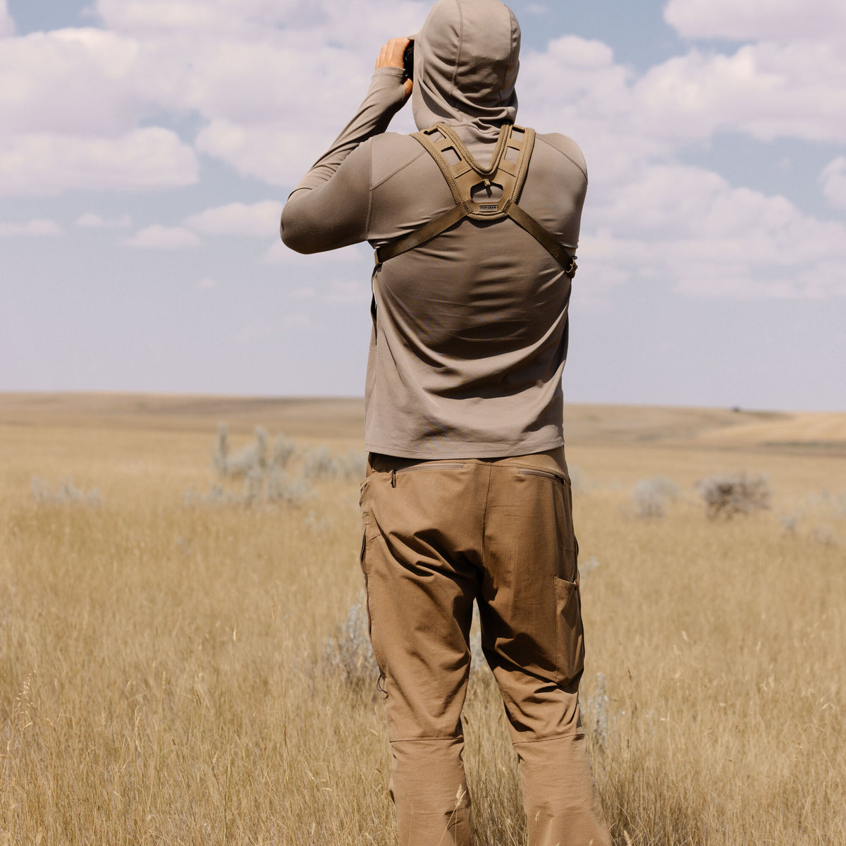 Person wearing a Walnut hooded shirt and brown cargo pants with an olive binocular harness, looking through binoculars in a grassy field.