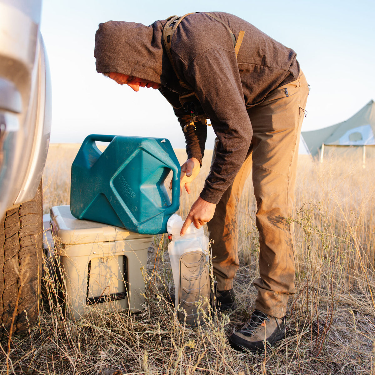 A person fills a clear Osprey hydration bladder from a teal Reliance water container resting on a tan cooler.