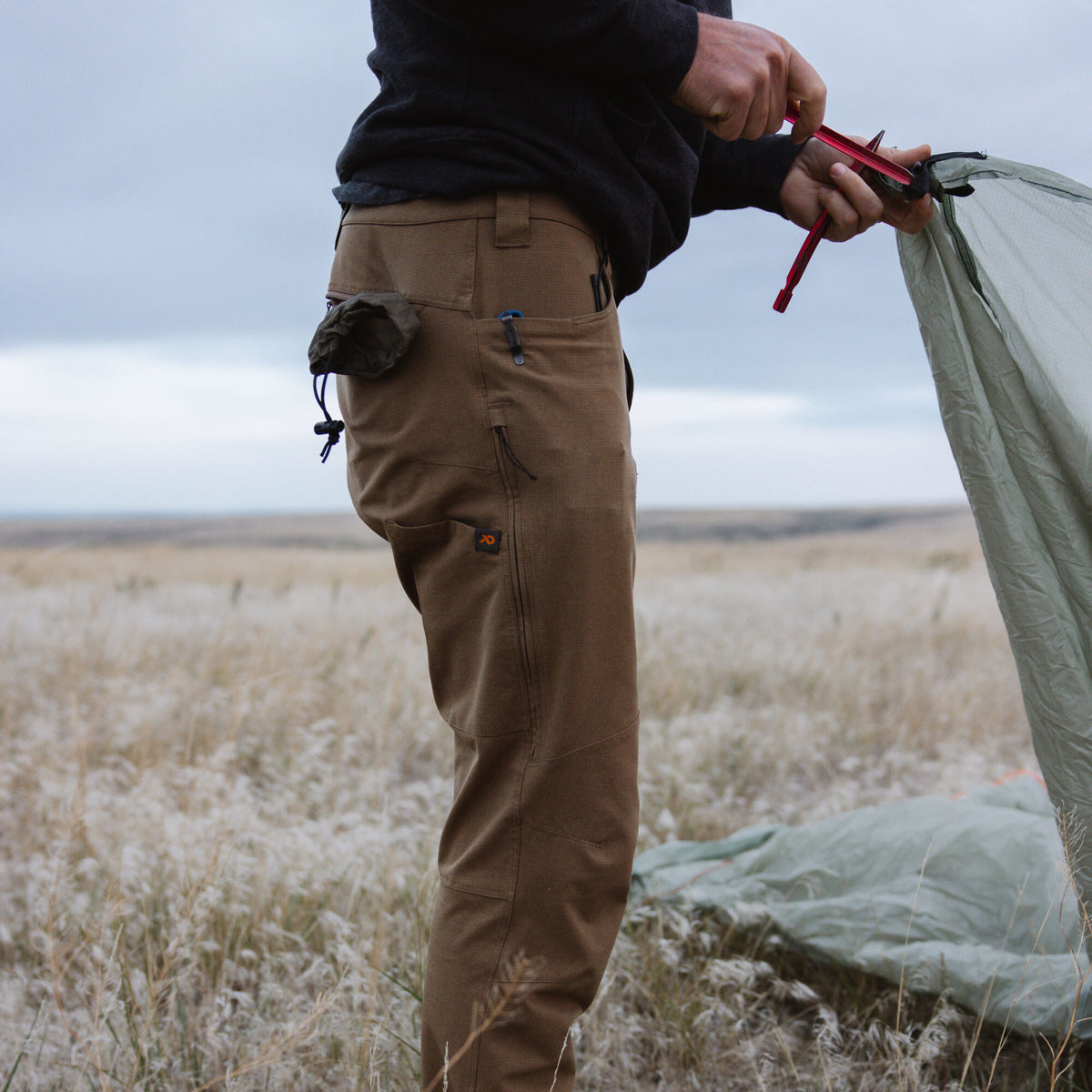 Brown outdoor pants featuring multiple pockets, an orange logo, and articulated knees, worn by a person setting up a tent.