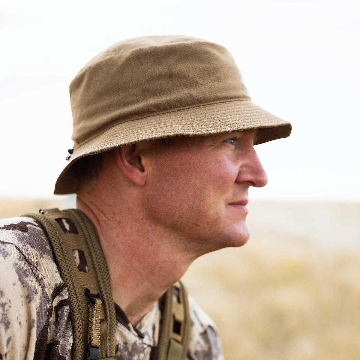 Tan boonie hat with a wide brim and subtle perforated texture, worn by a person in camouflage.