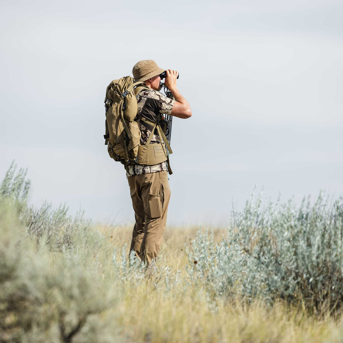 A man wearing a large tan hunting backpack with a bow, tan cargo pants, and a tan hat looks through binoculars.