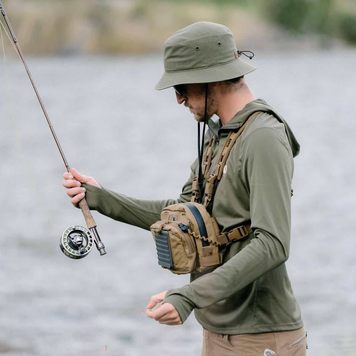 A man wears a tan fishing chest pack with black and grey features over an olive green long-sleeve shirt with thumbholes, and an olive green bucket hat, while fly fishing.