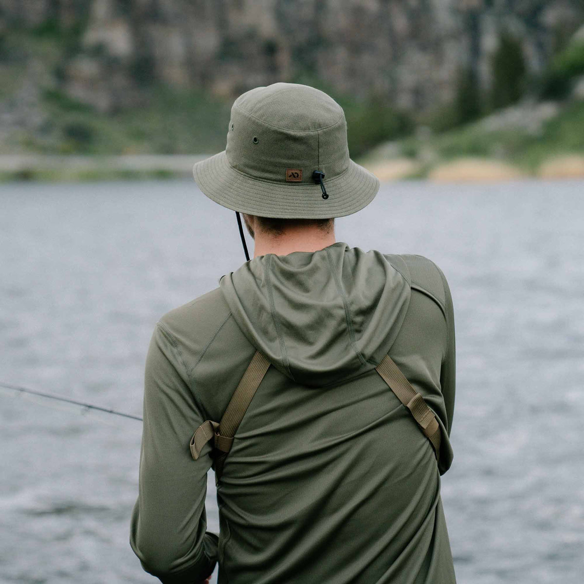 Olive green bucket hat with a wide brim, ventilation eyelets, chin strap with cord lock, and a small brown brand label.