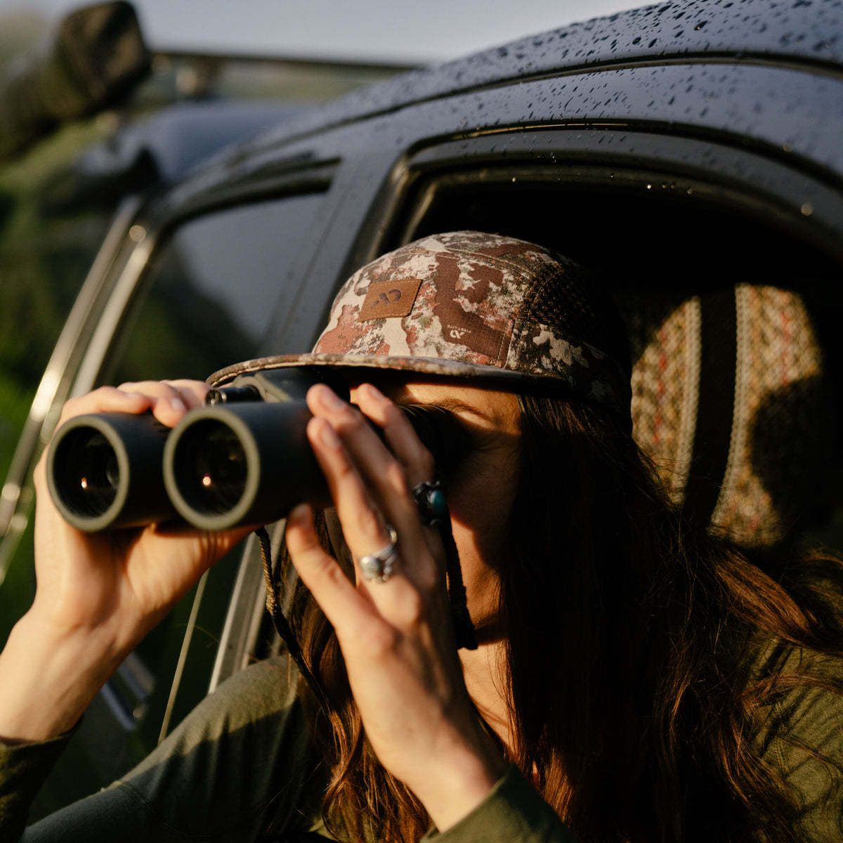 A woman wears a brown, beige, and green camouflage cap with a brown logo patch.