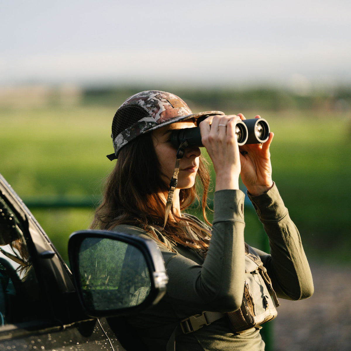 A woman wearing a camouflage mesh-back cap, olive long-sleeve shirt, and tan chest harness looks through black binoculars.
