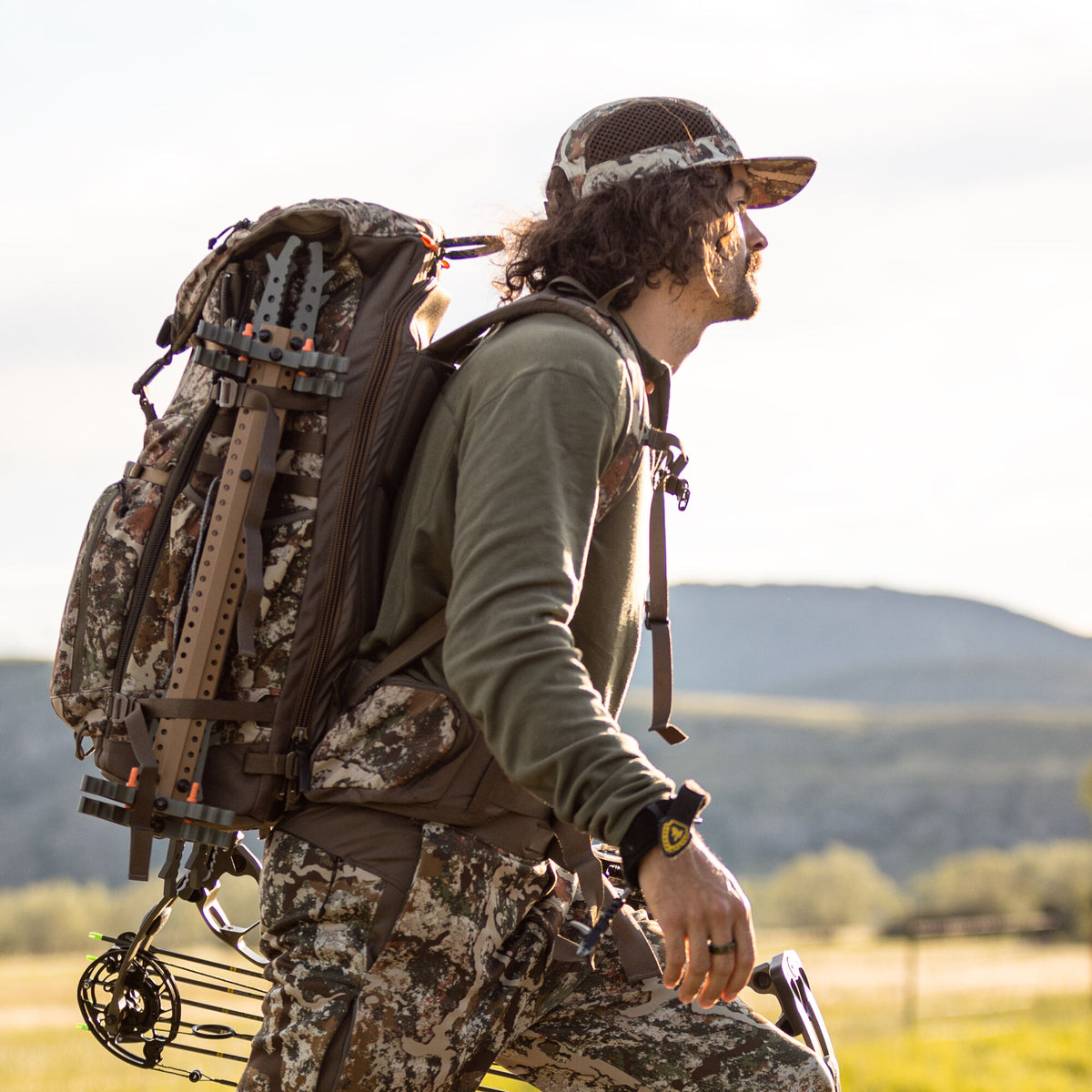 Camouflage hunting backpack system with an attached tan climbing stick and a black compound bow with green arrows.