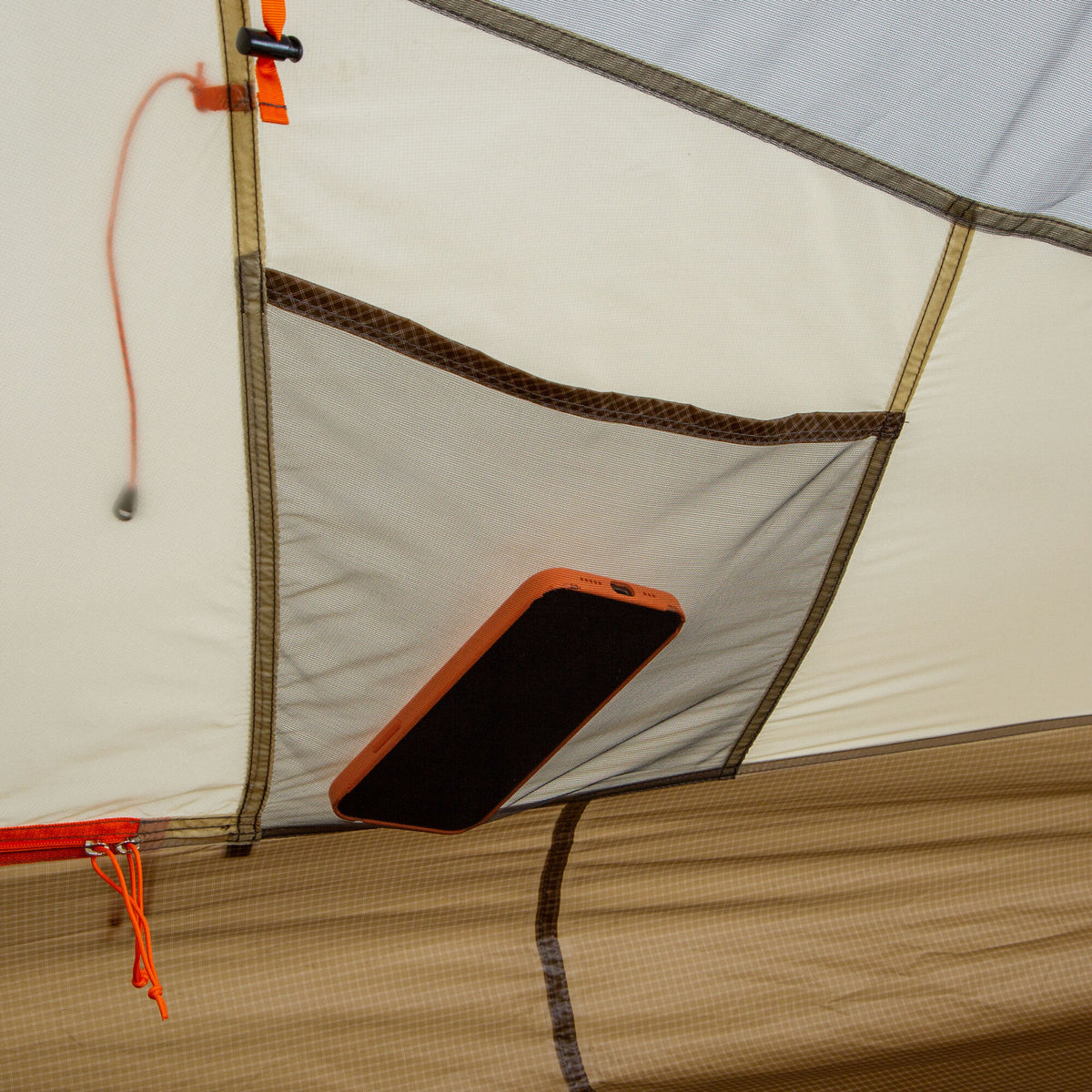 An orange phone sits in a dark mesh pocket inside a two-tone beige and tan tent, featuring an orange zipper pull.