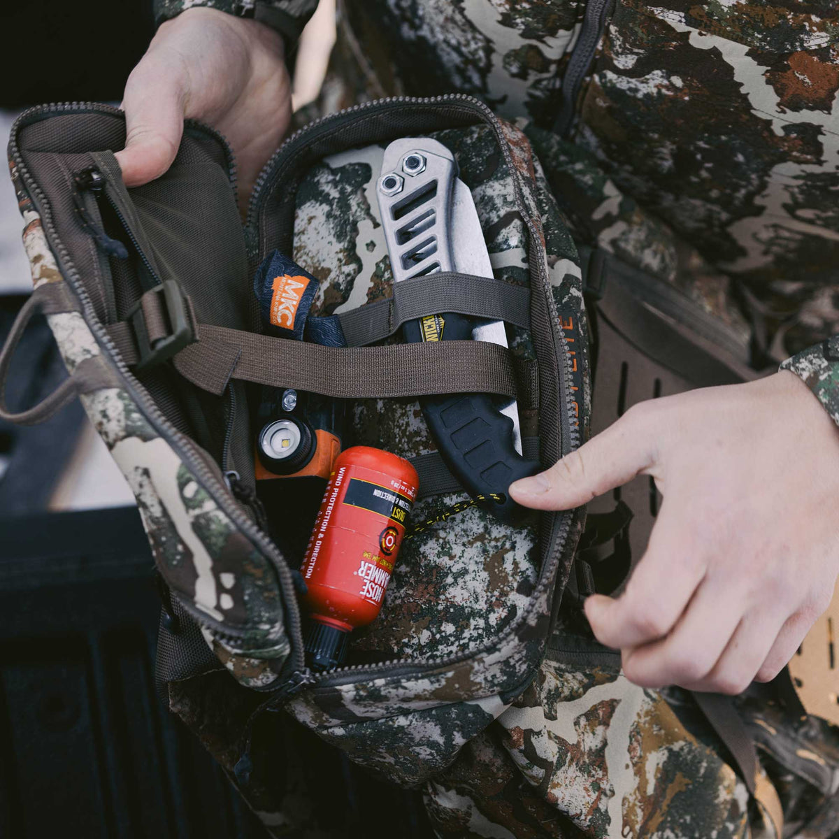 A person opens a camouflage hunting pouch, revealing a knife, headlamp, and red scent eliminator bottle secured within.