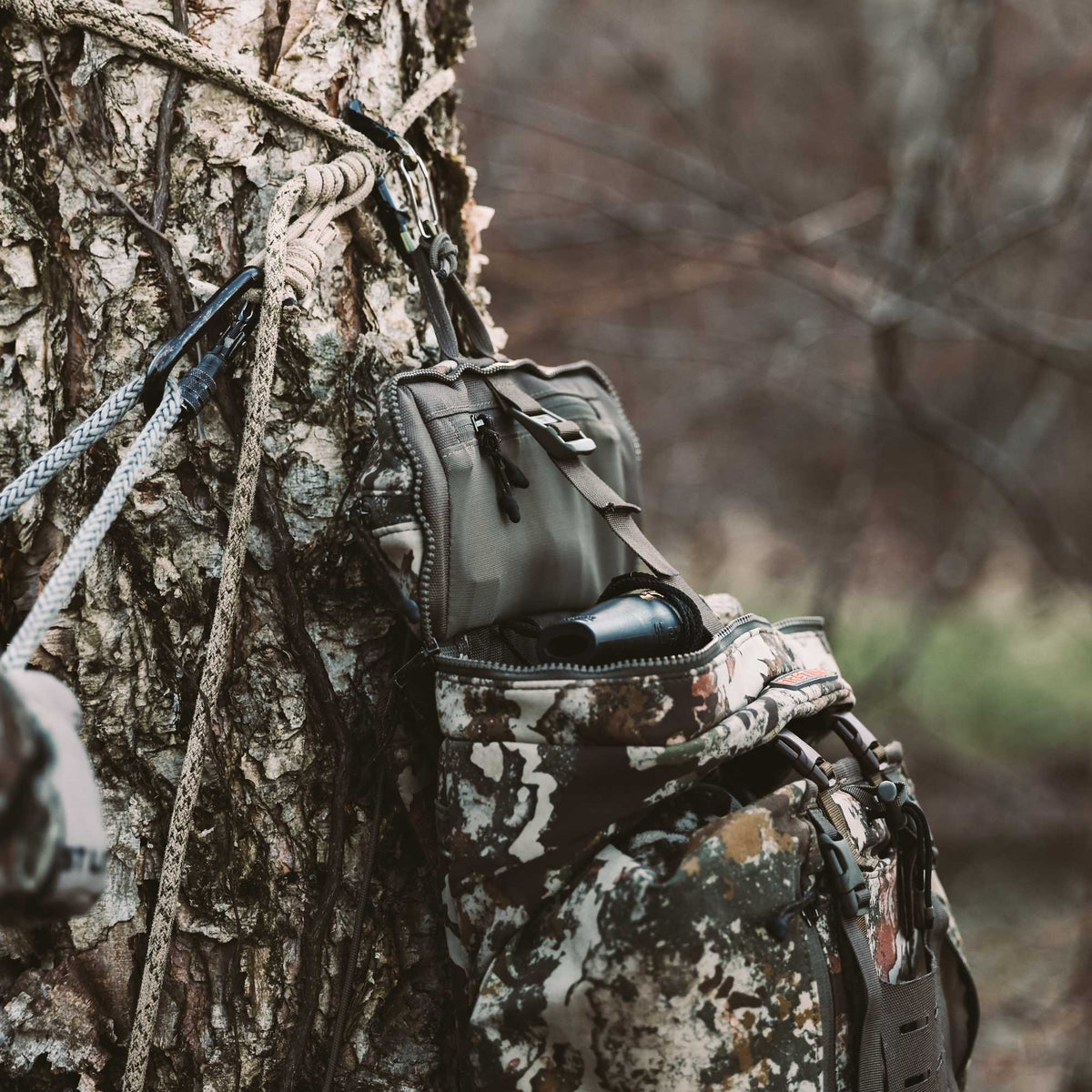 Camouflage hunting pack with an olive utility pouch unzipped to show a black call, suspended from ropes on a tree.
