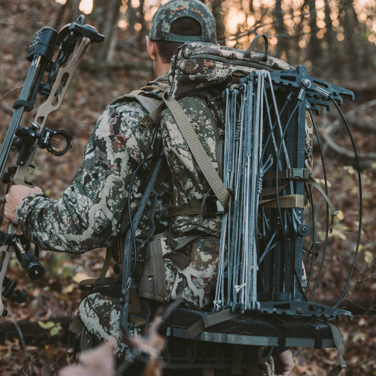 A hunter in camouflage carries a camo backpack, a bow, and black Shikar climbing sticks with ropes through a wooded area.