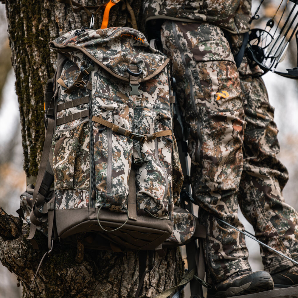 First Lite Fusion camouflage hunting backpack with visible logo, multiple pockets, compression straps, and dark brown base, hanging from a tree.