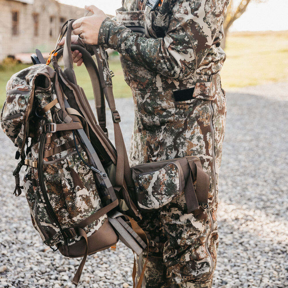 A person wearing a camouflage outfit holds a multi-tone camouflage hunting backpack with visible straps and pockets.