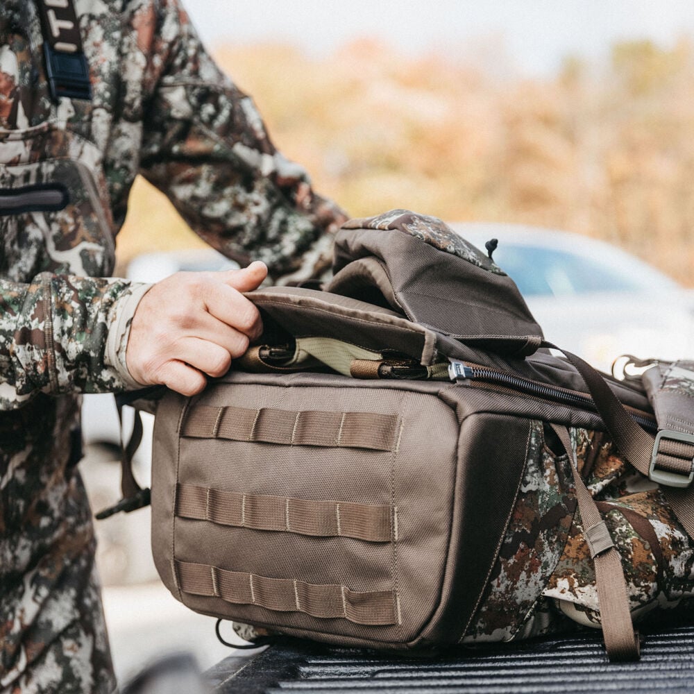 A person's hand opens a brown hunting pack with camouflage accents and MOLLE webbing.