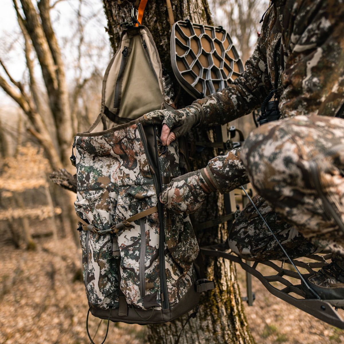 Camouflage hunting backpack with a vertical zipper being closed by a hunter's gloved hand, hanging from a tree.