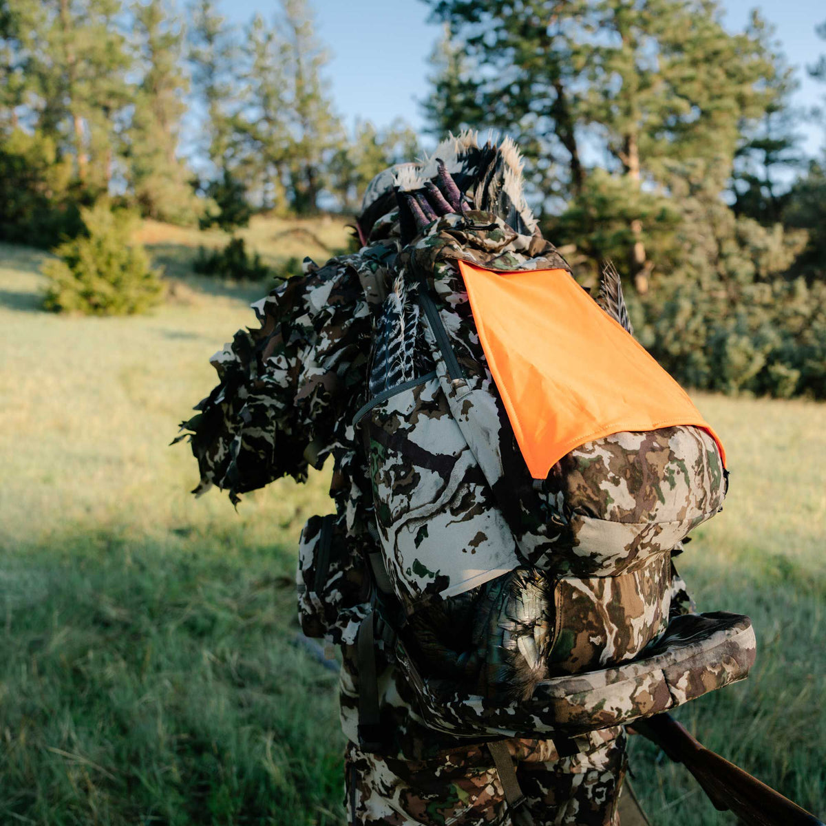 Hunter in a multi-color camouflage hunting outfit and backpack with a bright orange safety panel, turkey feathers, and a slung rifle.