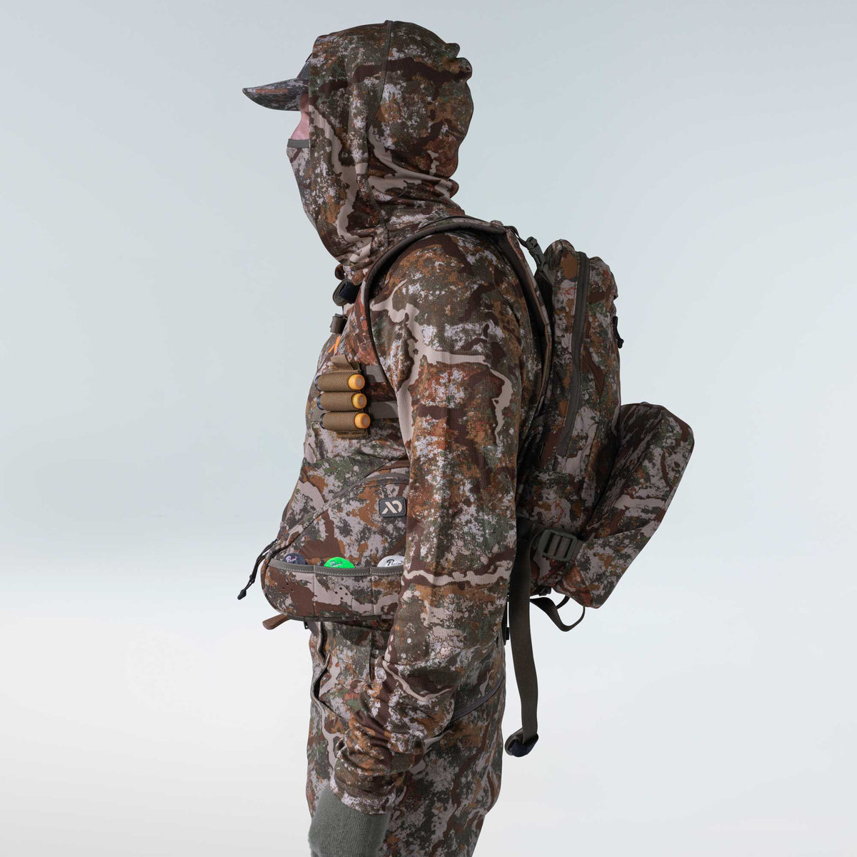 Full hunter's outfit in an earth-toned camouflage pattern, featuring a matching balaclava, cap, backpack, chest rig with three orange-capped cartridges, and a waist pouch.