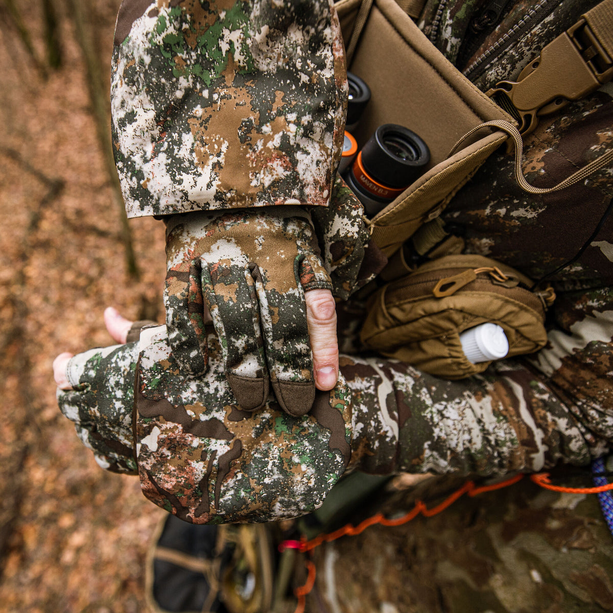 Person in multi-color camouflage jacket and gloves with exposed fingertips, wearing a tan chest rig holding black binoculars and a white bottle.