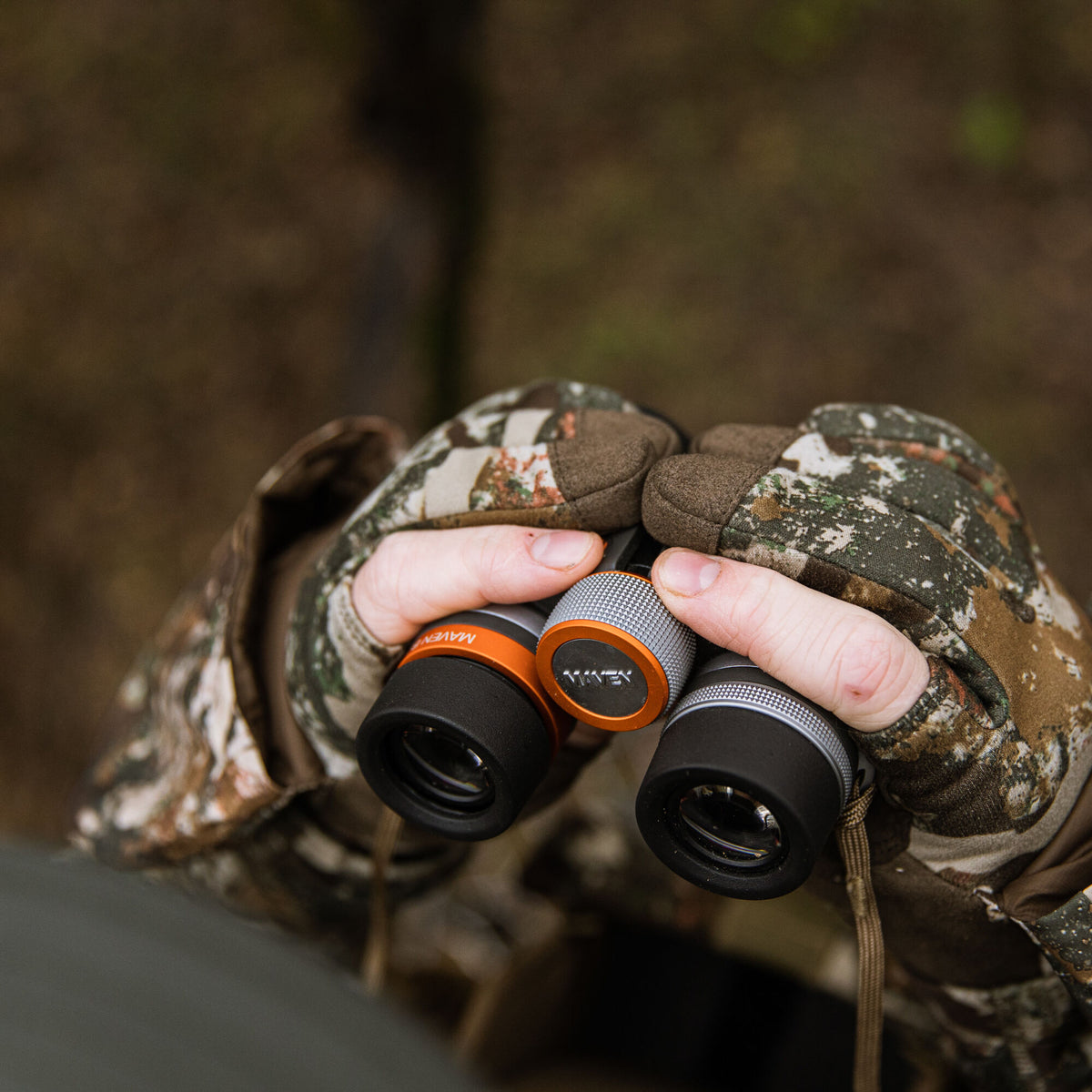 Hands in camouflage gloves hold black binoculars with orange rings, silver knurled focus rings, and a visible MAVEN logo.