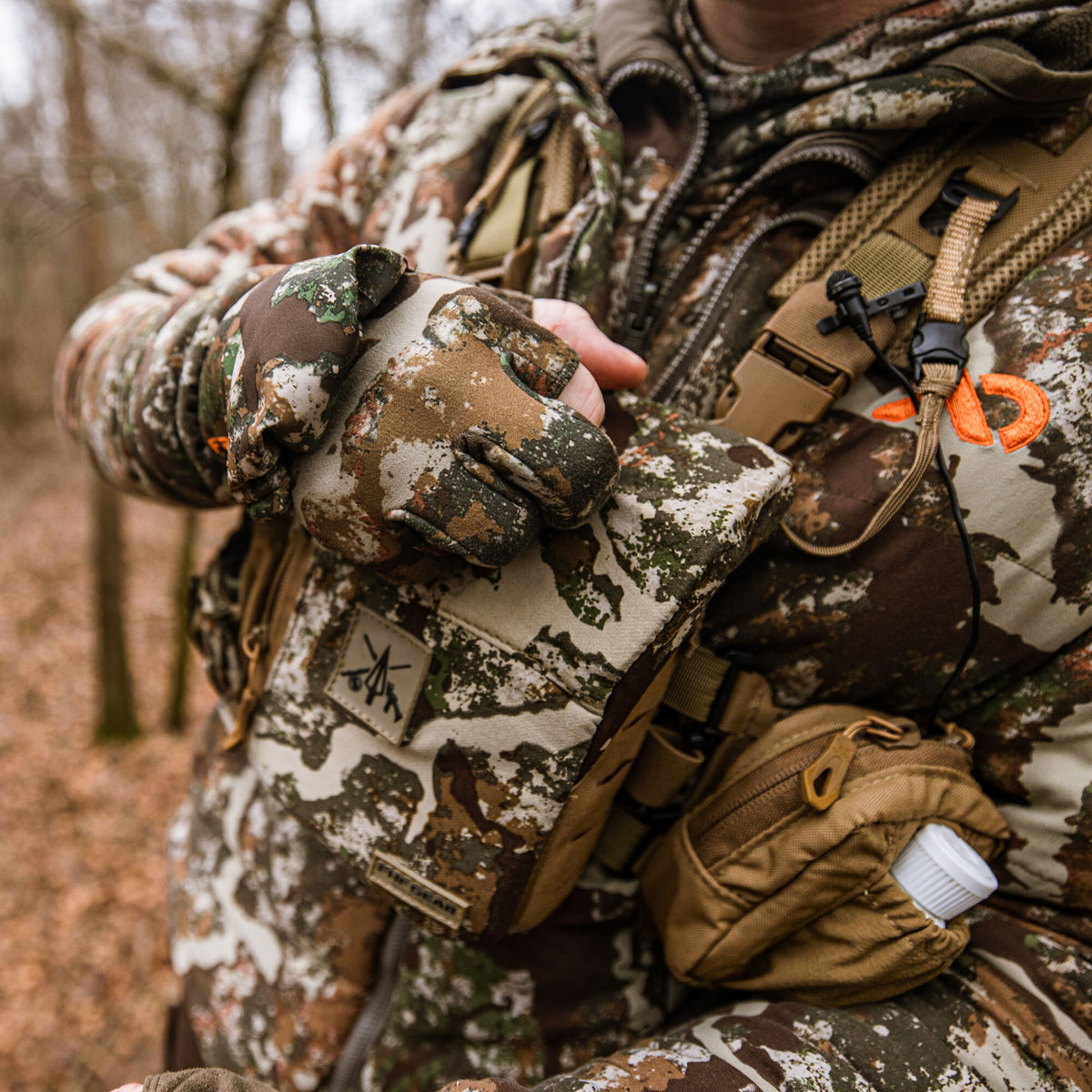 A person wears multi-tone camouflage hunting apparel, fingerless gloves, and a chest rig with a patch and a tan accessory pouch holding a white bottle.