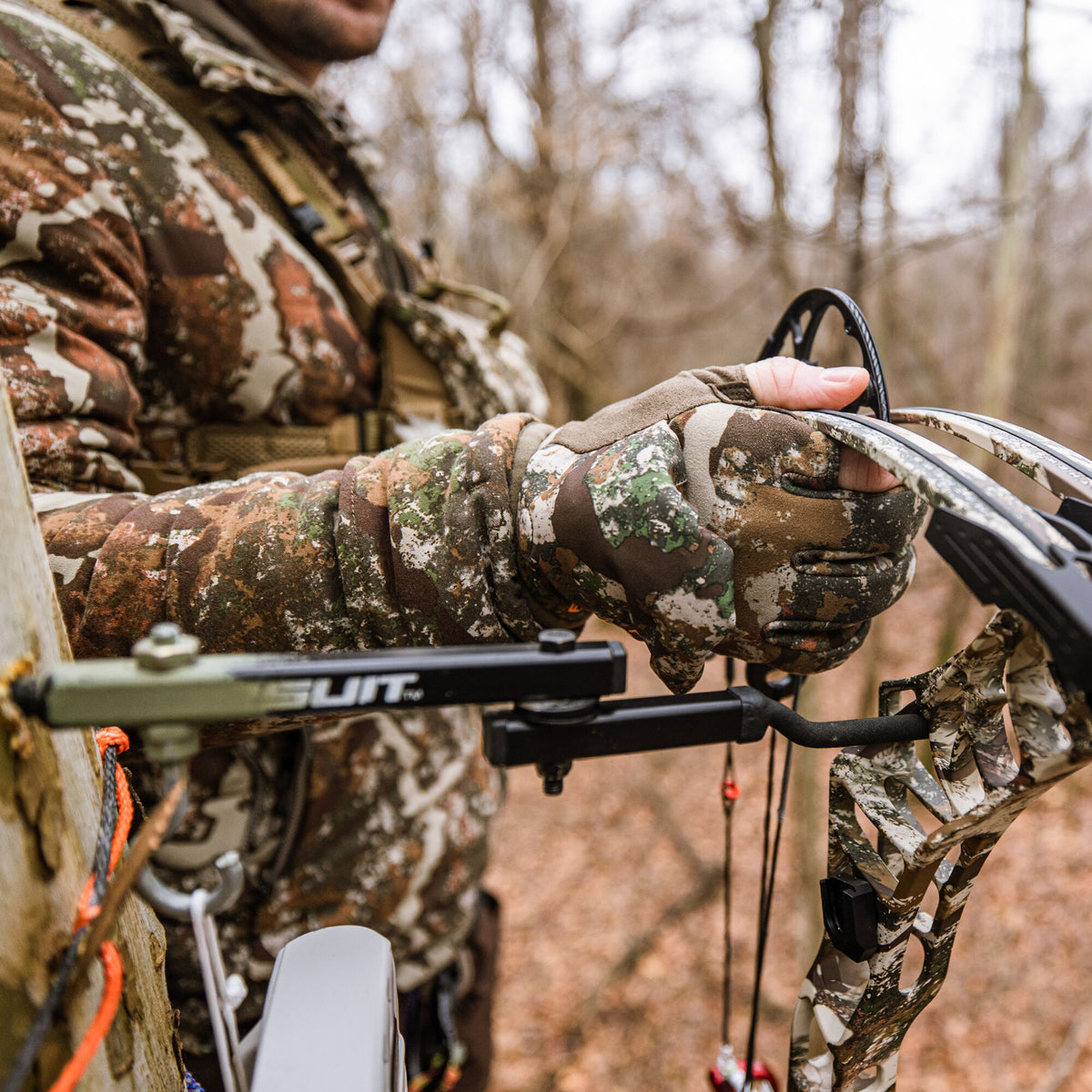 A hand in a brown, green, and white camouflage hunting glove holds a compound bow.